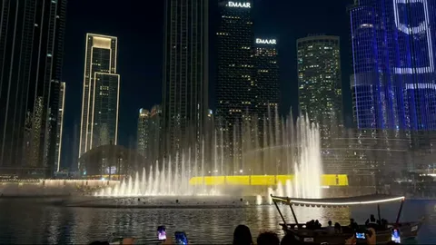 Dubai Fountain illuminated at night with skyline towers and a traditional boat in view