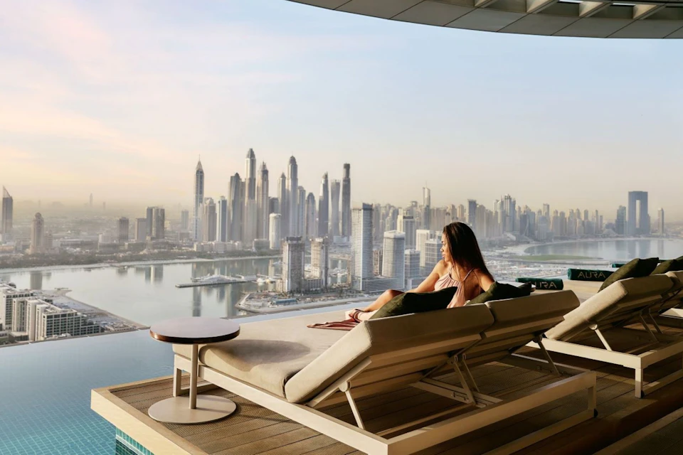 A woman relaxes on a lounge chair by an infinity pool, overlooking a cityscape with tall skyscrapers and a river.