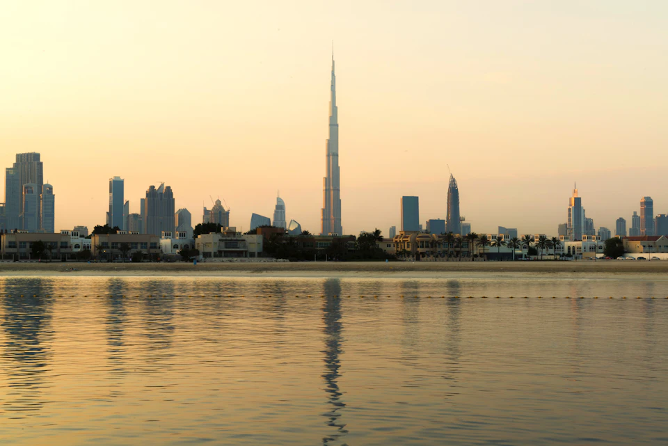 Skyline view of a city with various skyscrapers, including a central tall tower, reflecting on calm water during sunset.