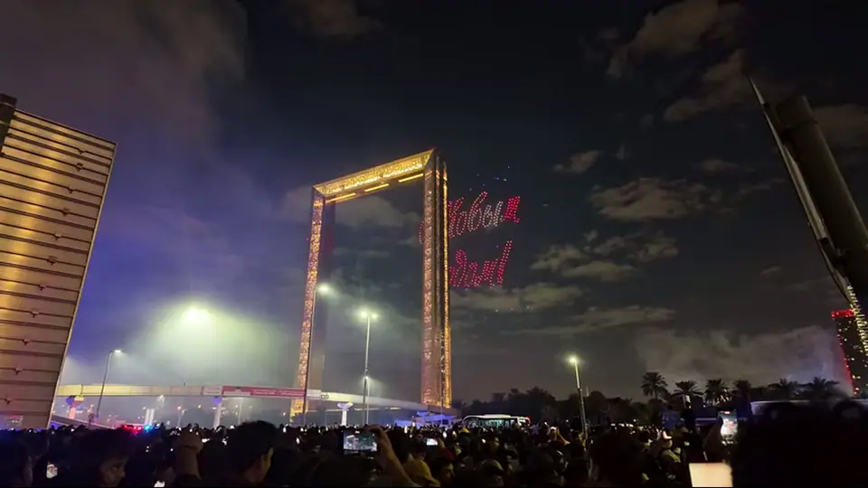 Crowd films fireworks exploding around Dubai Frame at night.