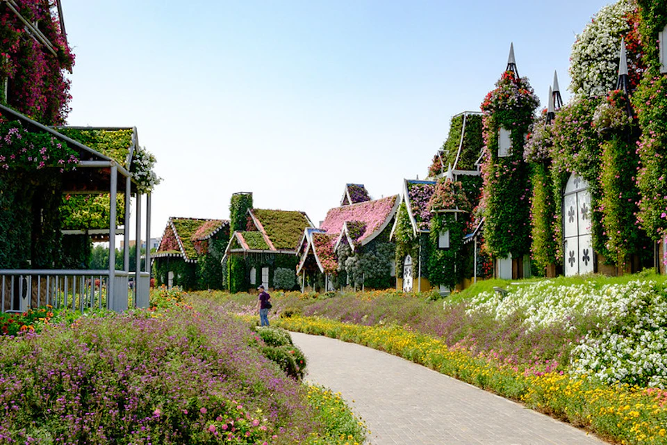A flower-lined path leads to houses fully covered in greenery and flowers under a clear sky; a person stands in the distance.