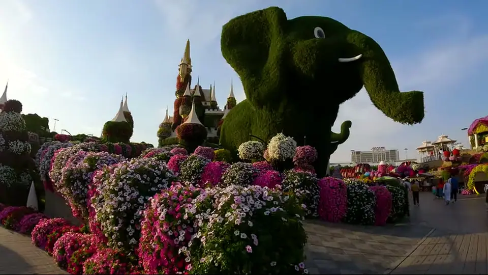 Huge floral elephant above flower beds and towers in warm light.