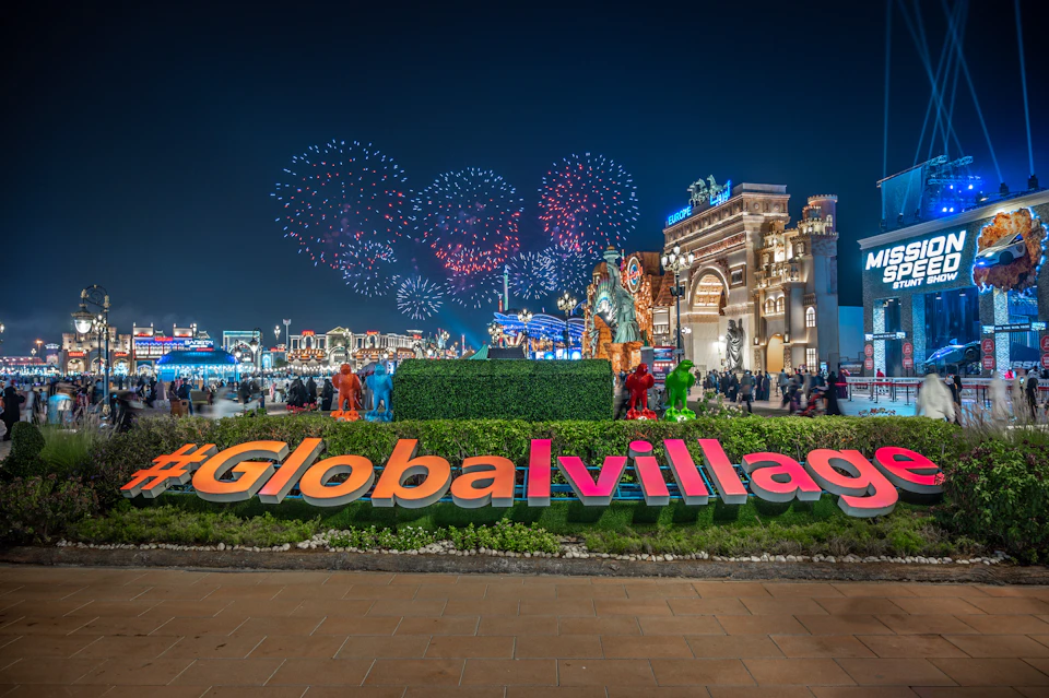 Night scene of #GlobalVillage with illuminated attractions, crowds of people, colorful fireworks, and a sign for "Mission Speed Stunt Show."