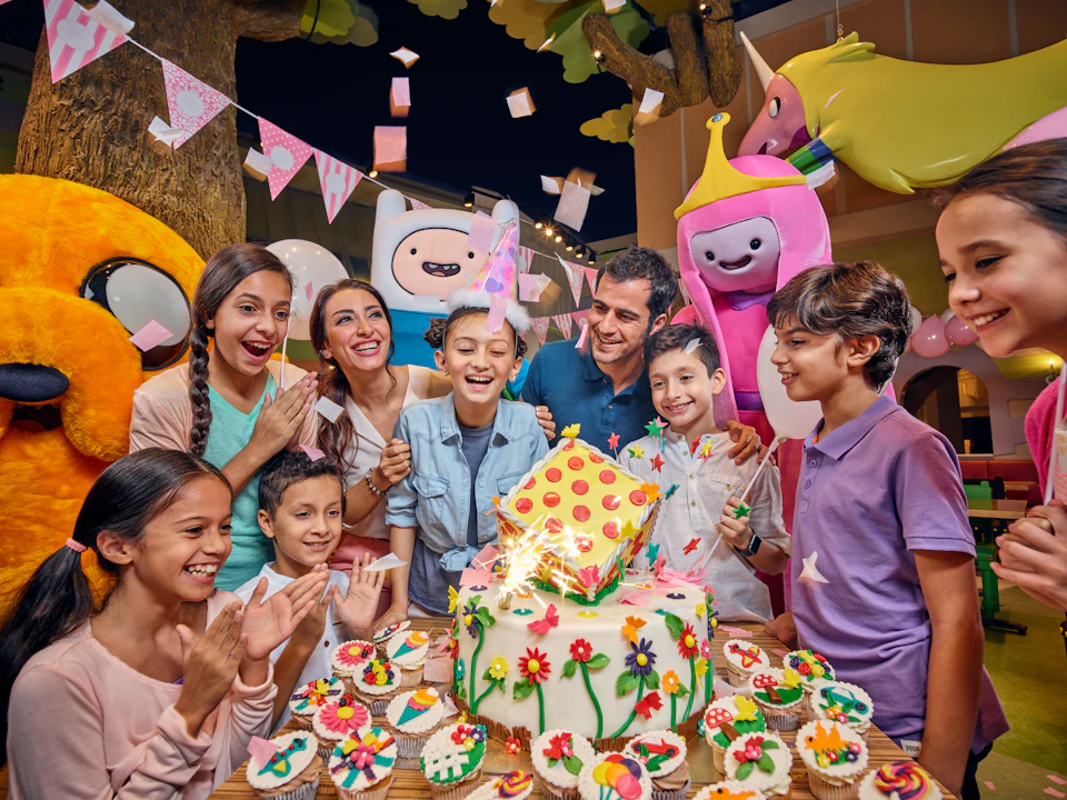 Group of children and adults celebrating a birthday with a colorful cake, cupcakes, decorations, and cartoon character mascots.