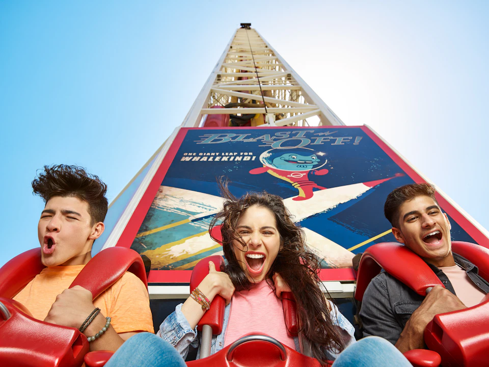 Three people strapped into a ride, looking excited and screaming, with clear blue sky and a "Blast Off!" sign behind them.