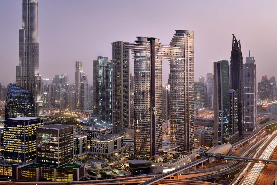 Panoramic view of a city skyline at dusk with modern skyscrapers, illuminated office buildings, and busy roads.