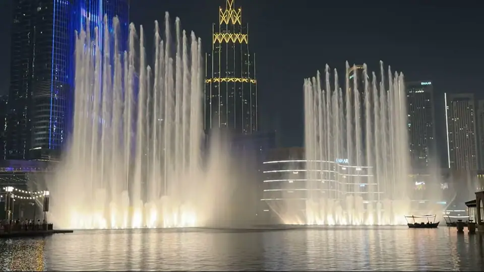 Освещенные струи фонтана стеной заполняют кадр, небоскребы позади ночью.