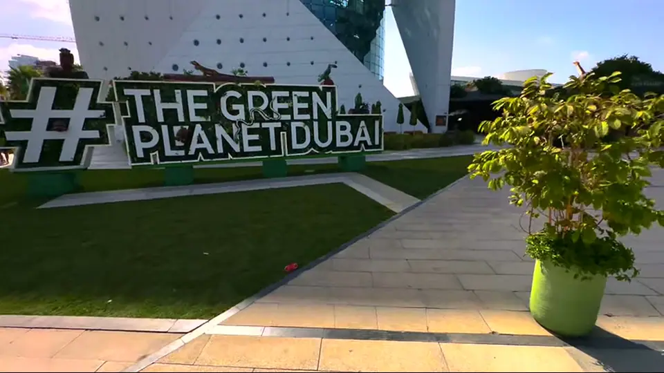 Знак The Green Planet Dubai на газоне у современного здания.